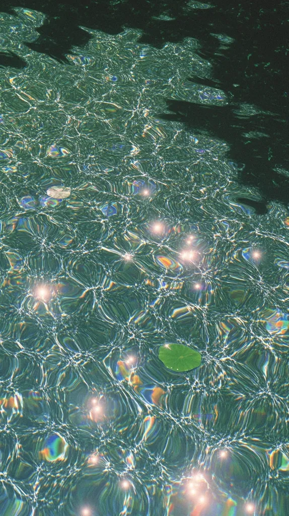 The image shows a close-up view of rippling water, possibly a swimming pool or calm lake.  Sunlight creates shimmering, iridescent reflections on the surface, with subtle color variations of teal and green.  A few small, dark objects, possibly leaves, float gently on the water. The overall effect is serene and visually captivating, emphasizing the interplay of light and water.