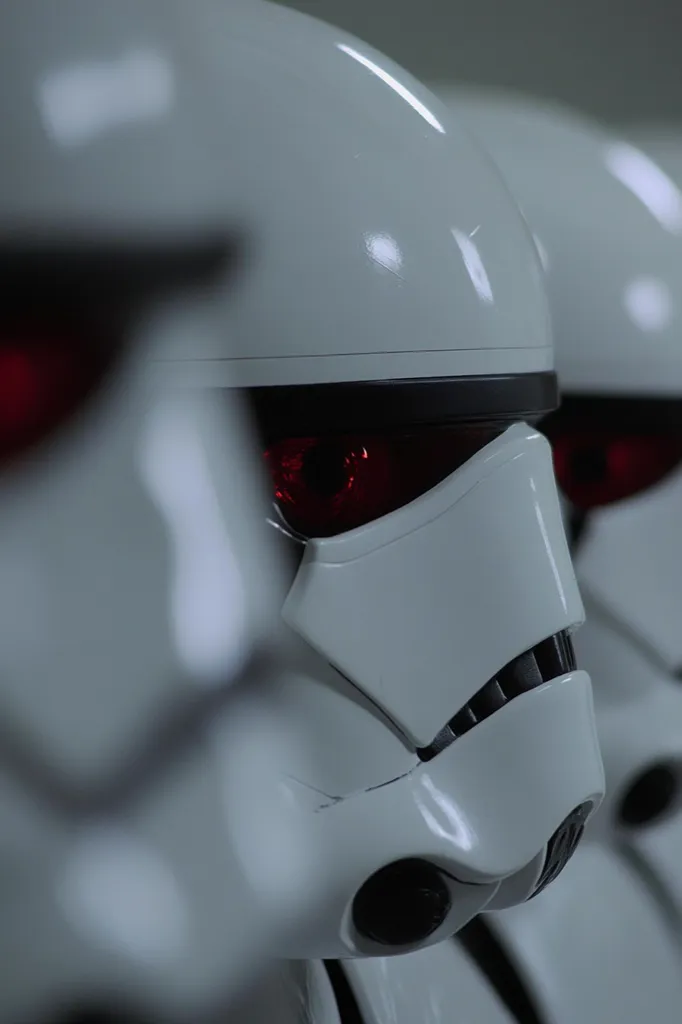 A close-up shot reveals a line of stormtrooper helmets, their gleaming white surfaces reflecting a dim light.  The focus is sharpest on the helmet in the foreground, showcasing the menacing red eye detail.  The depth of field blurs the helmets behind, creating a sense of a larger formation. The overall image is stark and imposing, hinting at a military presence.