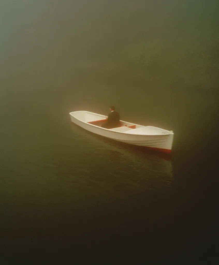 A lone figure sits in a small, white rowboat on a calm, foggy lake. The boat is centrally positioned in the frame, and the mist obscures the surrounding landscape. The overall mood is serene and somewhat mysterious, emphasizing solitude and the vastness of nature. The muted colors enhance the contemplative atmosphere.