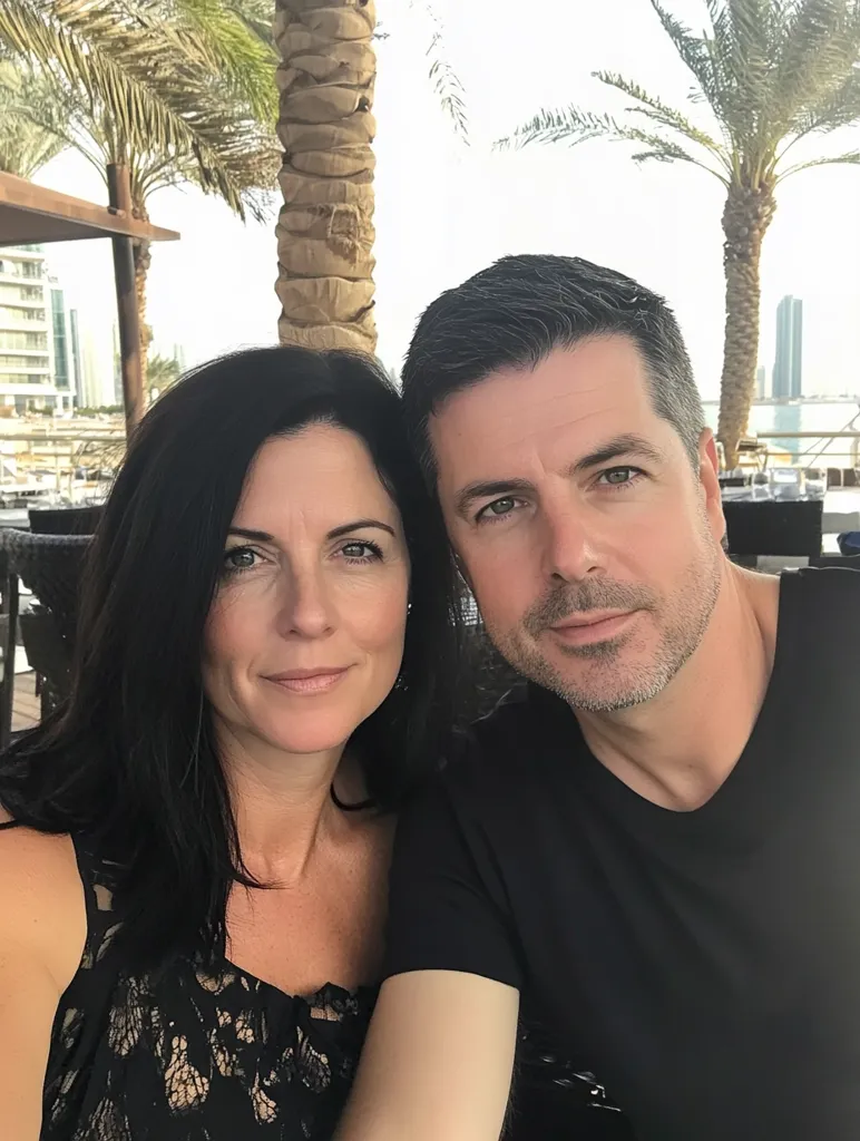A man and woman pose for a photo outdoors.  The woman, with long dark hair, wears a black lace top. The man, with short dark hair, wears a black t-shirt.  They are sitting close together, appearing relaxed and happy.  Palm trees and a waterfront cityscape are visible in the background.  The setting suggests a vacation or a pleasant day out.