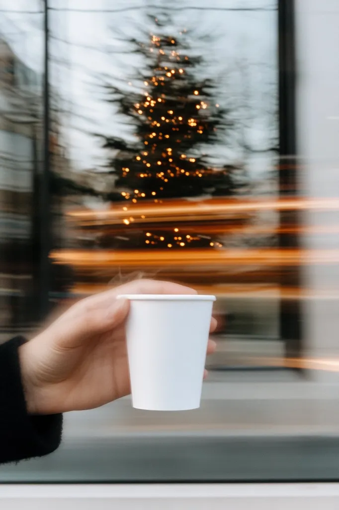 A hand holds a steaming white paper cup against a blurred background.  A brightly lit Christmas tree is visible through a window, its lights creating a warm glow.  The motion blur suggests movement, perhaps from a vehicle passing by. The overall image evokes a feeling of cozy warmth during the holiday season.