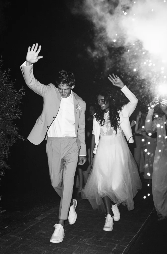 A black and white photo captures a newly married couple exiting their reception.  The groom, in a light-colored suit and white sneakers, waves to guests.  The bride, in a flowing tulle dress and white sneakers, smiles radiantly.  Sparklers create a celebratory atmosphere in the background as the couple joyfully makes their exit.