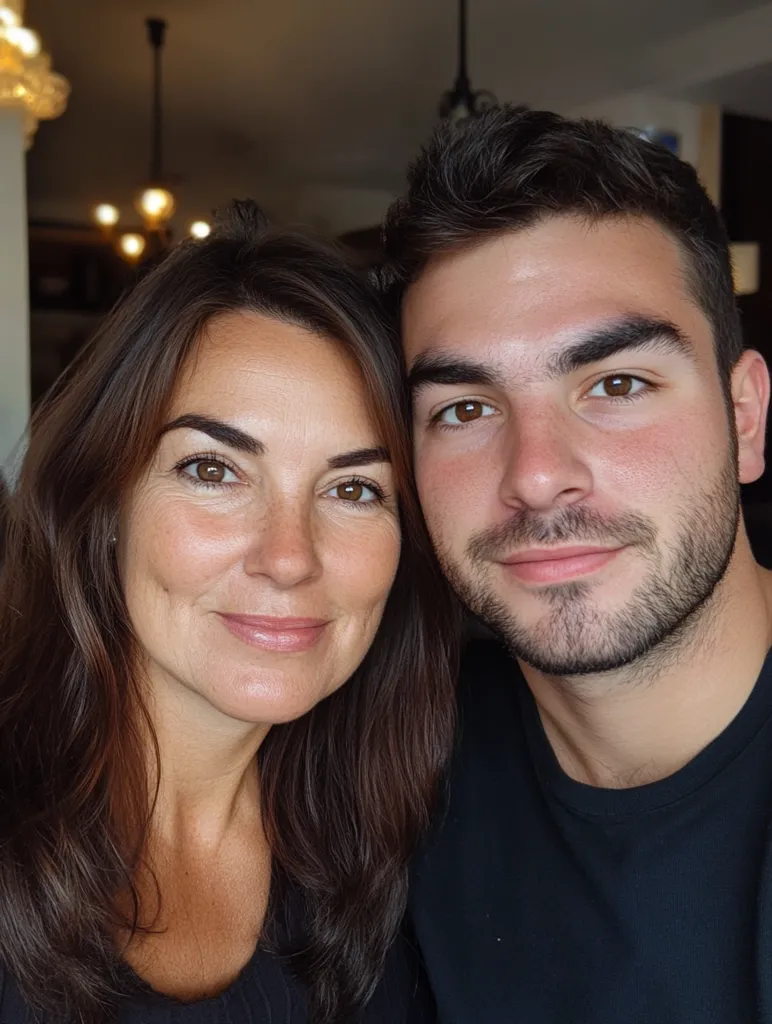 A close-up shot features a woman and a young man, likely mother and son, posing for a selfie. The woman, with long brown hair, smiles gently, her face showing warmth and maturity. The man, with short brown hair and a beard, has a softer expression, his gaze directed slightly away from the camera.  Both are dressed casually in dark clothing. The background is softly blurred, suggesting an indoor setting. The image conveys a sense of intimacy and familial connection.