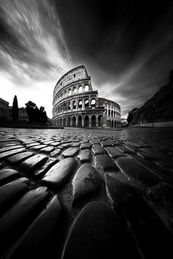 A dramatic black and white photograph captures the Colosseum in Rome.  The ancient amphitheater's curved structure dominates the frame, set against a sky of swirling clouds.  The foreground showcases a textured cobblestone street, leading directly to the iconic landmark.  The contrast between the dark stones and the light sky creates a powerful, timeless image.