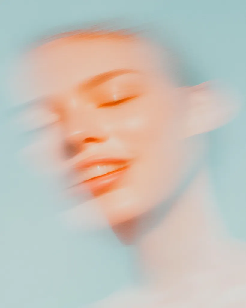 A dreamy, soft-focus image depicts a woman's face, eyes gently closed, exuding serenity.  Her skin is subtly highlighted with a warm, peachy tone against a cool, pale blue background. The blurred effect creates a sense of ethereal beauty and tranquility, emphasizing the smooth texture of her skin and the relaxed curve of her lips.
