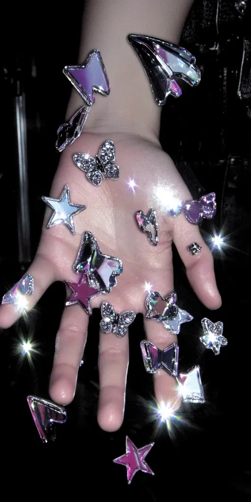 A hand is adorned with iridescent, jewel-toned charms shaped like butterflies, stars, and paper airplanes.  The charms are scattered across the hand and fingers, catching the light and creating sparkling highlights. The overall aesthetic is dark, glamorous, and slightly edgy.  The image evokes a sense of fantasy and whimsy.
