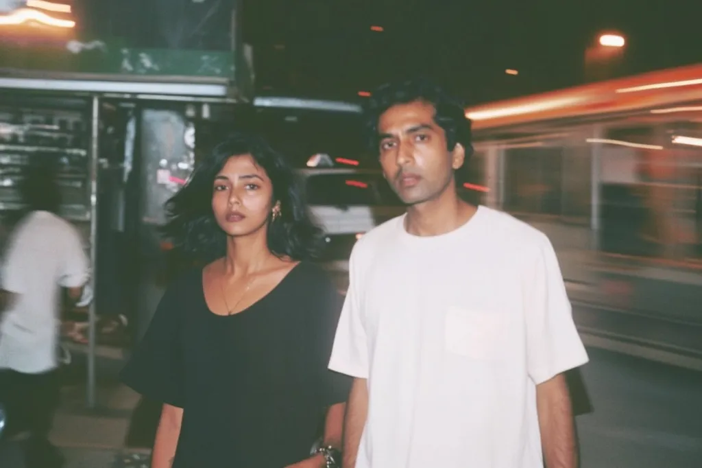 A photo of a young couple standing on a city street at night.  The woman wears a black shirt and the man a white t-shirt.  Blurred lights from vehicles and street signs create a nighttime urban backdrop. The image has a grainy, slightly out-of-focus quality, suggestive of a film photograph.  The couple appears to be walking or pausing in their journey.