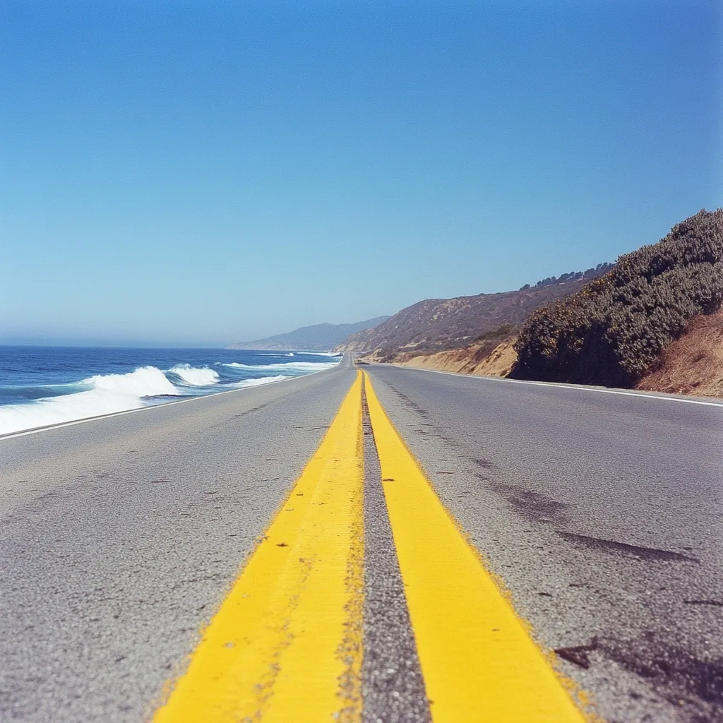 A scenic coastal highway stretches towards the horizon under a clear blue sky.  The asphalt road, marked by vibrant yellow double lines, leads to a tranquil ocean with gentle waves lapping the shore.  Dry, brown hills and sparse vegetation line the roadside, creating a beautiful contrast against the deep blue of the sea.  The perspective emphasizes the road's journey and the open space ahead.