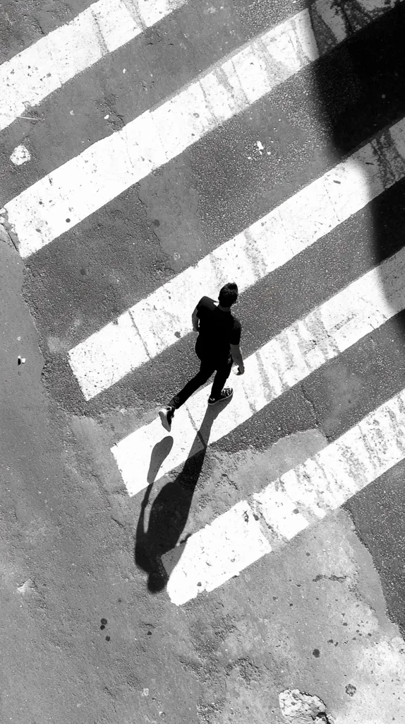 High-angle, black and white shot of a person walking across a pedestrian crossing. The individual is dressed in dark clothing, and their shadow is cast prominently on the pavement. The crossing's white stripes contrast sharply against the dark asphalt, creating a strong visual pattern. The overall scene is simple yet impactful, emphasizing the contrast between light and shadow and the solitary figure in motion.