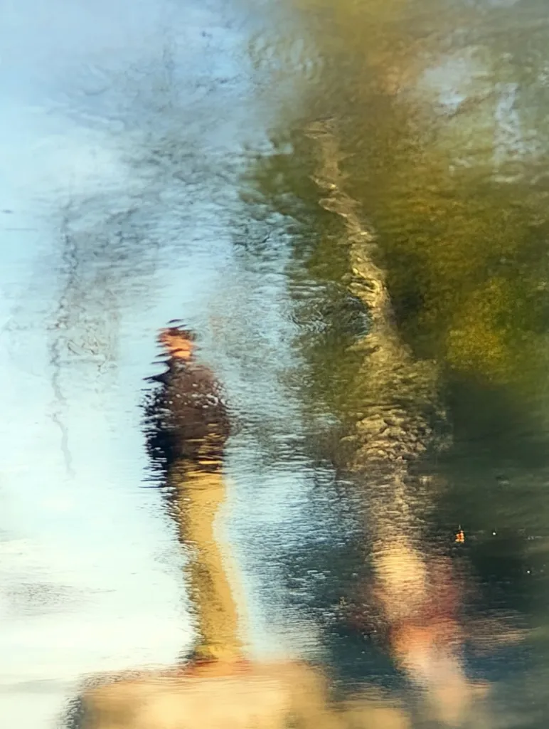 An abstract image showing the distorted reflection of a person standing near a tree in a puddle of water.  The reflection is blurred and fragmented, with the water's surface creating a wavy, textured effect. The colors are muted and blend, creating a dreamlike atmosphere. The overall impression is one of fluidity and impermanence.
