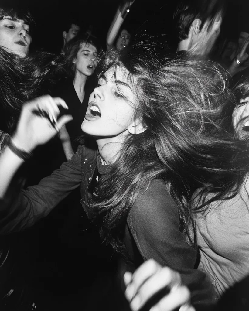 A black and white photograph captures the energy of a crowded concert. A young woman with long, flowing hair is at the center, her face a mask of exhilaration as she's caught in the throng. Surrounding her, other concertgoers are visible, faces blurred by motion and low light. The overall mood is one of intense excitement and movement, characteristic of a live music performance. The image's grain and contrast emphasize the chaotic yet captivating atmosphere.