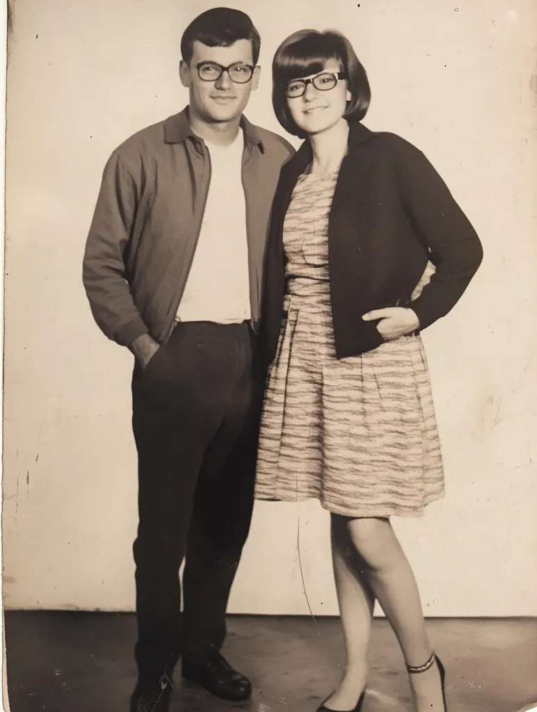 A black and white photograph shows a young couple posing for a portrait.  The man, wearing glasses and a zip-up jacket, stands slightly behind the woman. She is wearing a short, striped dress and a dark cardigan, also sporting glasses.  Both are dressed in the style of the 1960s. The image has a slightly faded, vintage quality.