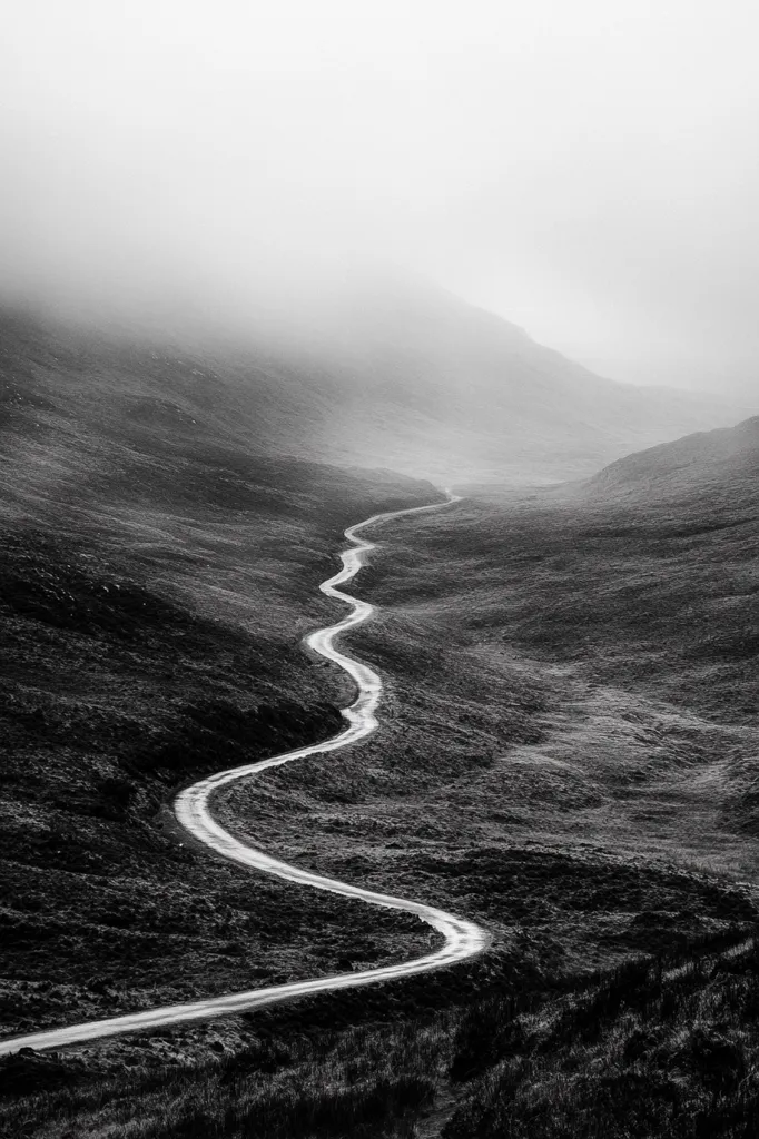 A winding road snakes through a misty, mountainous landscape.  The monochrome image emphasizes the road's curves as it disappears into the fog-shrouded hills. The scene evokes a sense of journey and the unknown, with the gray tones enhancing the atmospheric perspective.  The road is the focal point, drawing the eye through the desolate yet beautiful terrain.