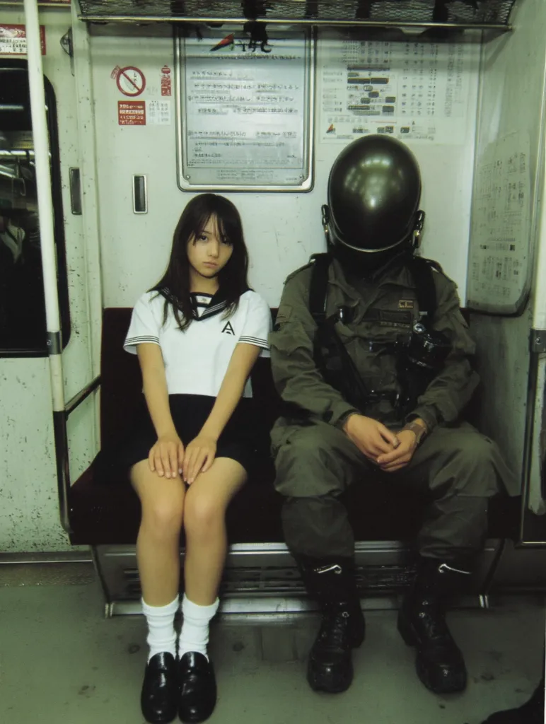 A young girl in a school uniform sits beside a person in military gear wearing a helmet on a public transit train.  The girl has long dark hair and is wearing white socks and black shoes. The soldier-like figure sits rigidly, hands clasped. The scene is dimly lit, emphasizing the contrast between the girl's youth and the soldier's serious demeanor.  The setting appears to be a Japanese train car.