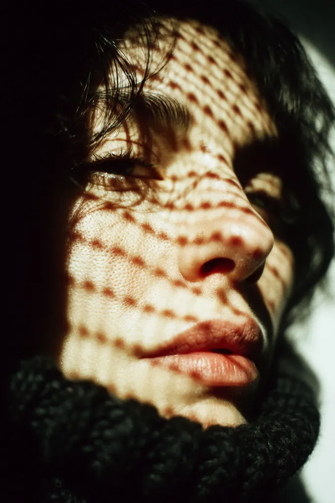 Close-up of a person's face, partially obscured by shadows cast from a patterned surface.  The dark hair frames a portion of the face, illuminated by dappled sunlight creating a reticulated pattern on the skin.  The individual's eyes are partially closed, and they wear a dark, textured knit collar. The overall mood is one of quiet contemplation.