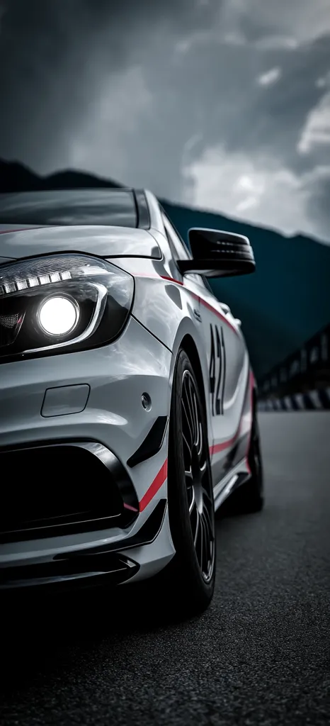 A close-up shot of a silver Mercedes-Benz A-Class, possibly a performance variant, parked on a dark asphalt road.  The car features red and black racing stripes along its side and the number 422 is visible. The headlight is illuminated, and the overall mood is dramatic due to the dark, cloudy sky in the background. The focus is sharply on the front of the vehicle.