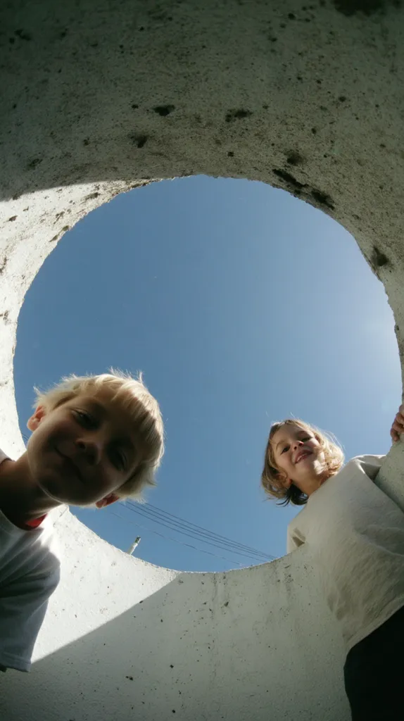 Two children peek out from the bottom of a large, round concrete pipe.  The clear blue sky is visible through the opening above them.  The children appear to be looking up and are smiling. The concrete is light gray and shows signs of age and wear. The scene is bright and sunny.