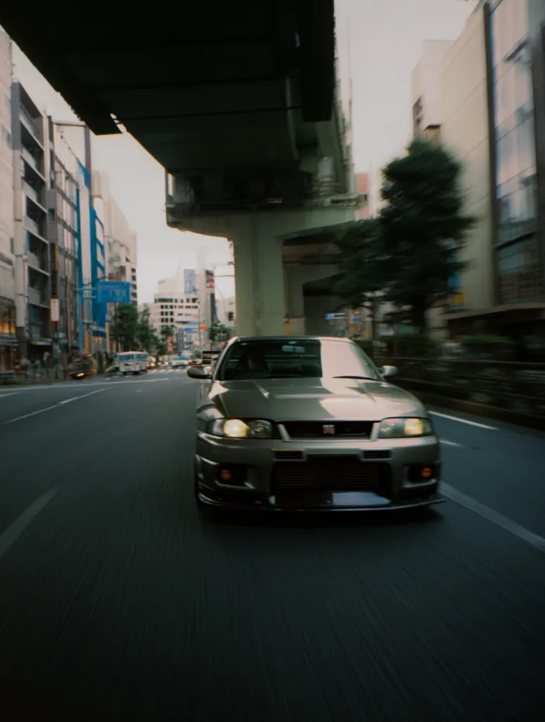 A metallic gray Nissan Skyline R34 GT-R drives down a city street in Japan. The car is in the foreground, with blurred buildings and an overpass visible in the background.  The city appears busy, but the focus is on the powerful sports car speeding along the road.  The image has a cinematic feel, reminiscent of a street racing scene.