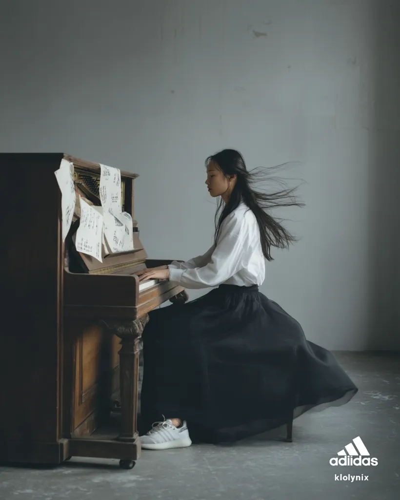 A young woman with long black hair flowing behind her sits at an antique upright piano.  She wears a white shirt and a long, black skirt, her Adidas sneakers visible beneath. Sheet music rests on the piano's music rack as she plays, creating a serene and artistic image.  The background is a muted grey, emphasizing the woman and the piano.