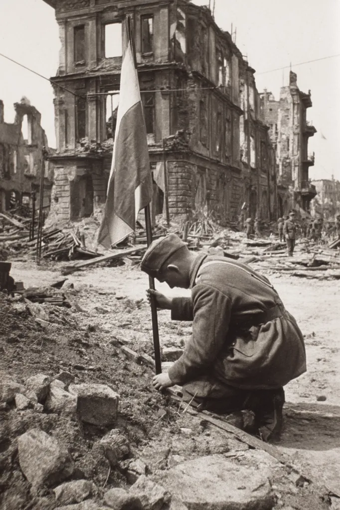 A black and white photo depicts a soldier kneeling in the rubble of a bombed-out city. He carefully plants a flagpole bearing a Polish flag into the ground amidst the devastation.  The background shows heavily damaged buildings, a stark testament to the destruction of war.  Other soldiers are visible in the distance. The image evokes a sense of somber resilience and the reclaiming of territory.