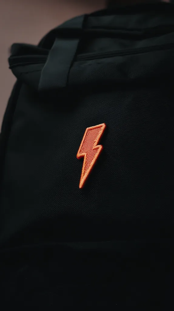 Close-up view of a black backpack with an orange embroidered lightning bolt patch. The backpack appears to be made of a durable, textured fabric. The lightning bolt is centrally positioned and is a vibrant contrast to the dark background.  The image focuses on the detail of the patch and the texture of the bag.