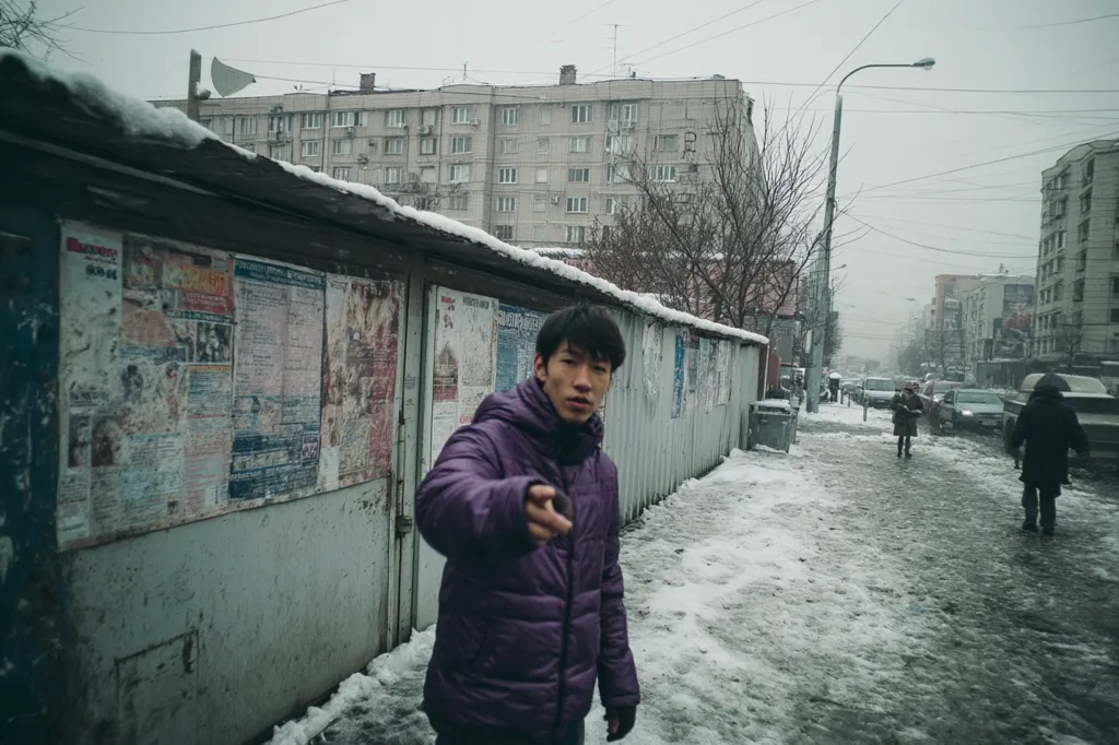 A young man in a purple puffer jacket stands on a snowy street in a city.  He's positioned in front of a weathered metal shed covered in faded posters.  The background shows apartment buildings and other people walking in the light snowfall. The overall atmosphere is muted and somewhat bleak.