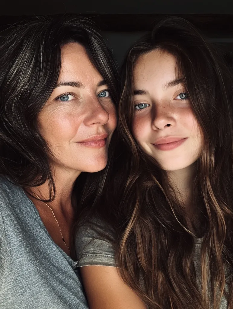 A close-up shot captures a mother and daughter, their faces close together.  The mother, with long, dark hair streaked with gray, smiles gently.  Her daughter, with long, brown hair, mirrors her expression. Both have fair skin and light eyes. They appear relaxed and affectionate, showcasing a tender moment of closeness.  The image is softly lit, emphasizing their features and creating a warm, intimate atmosphere.