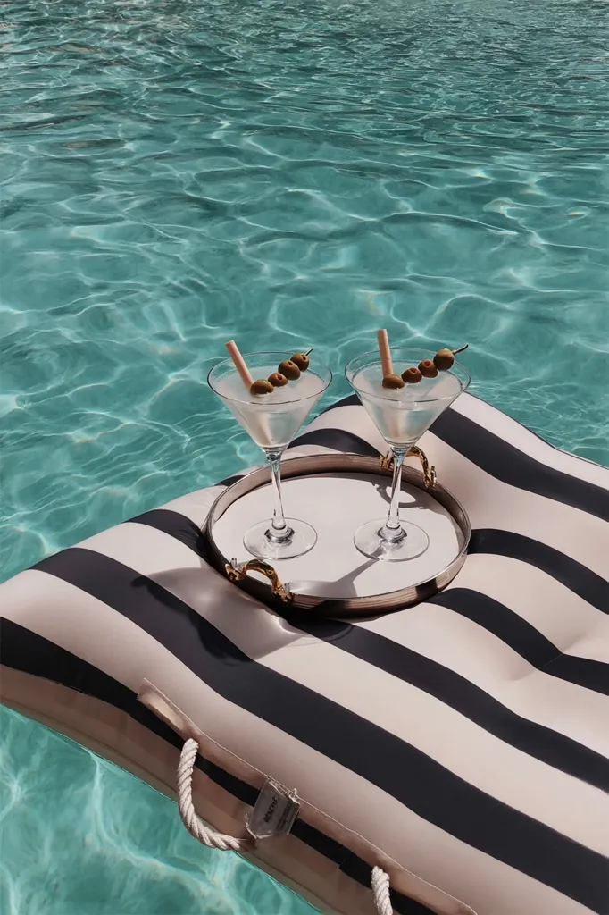 Two martinis garnished with olives rest on a round tray atop a black and beige striped inflatable pool float.  The float is partially submerged in a sparkling turquoise swimming pool.  The scene evokes a luxurious and relaxing poolside atmosphere.  The image is clean and aesthetically pleasing, emphasizing summer vibes and sophisticated leisure.