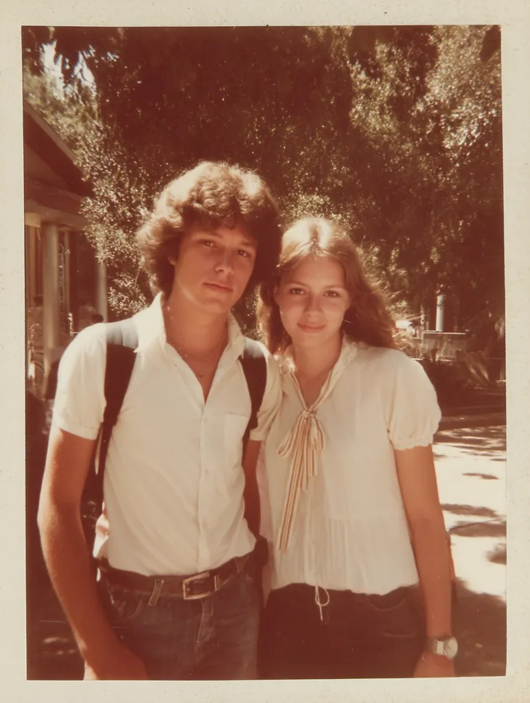 A vintage photograph shows a young couple standing outdoors on a sunny day.  The boy, with curly brown hair, wears a white button-down shirt and jeans, and a backpack. The girl, with long brown hair, sports a cream-colored blouse and dark jeans.  They stand close, suggesting a romantic relationship, against a backdrop of trees and a partially visible house.  The image has a warm, nostalgic quality, characteristic of a 1970s snapshot.