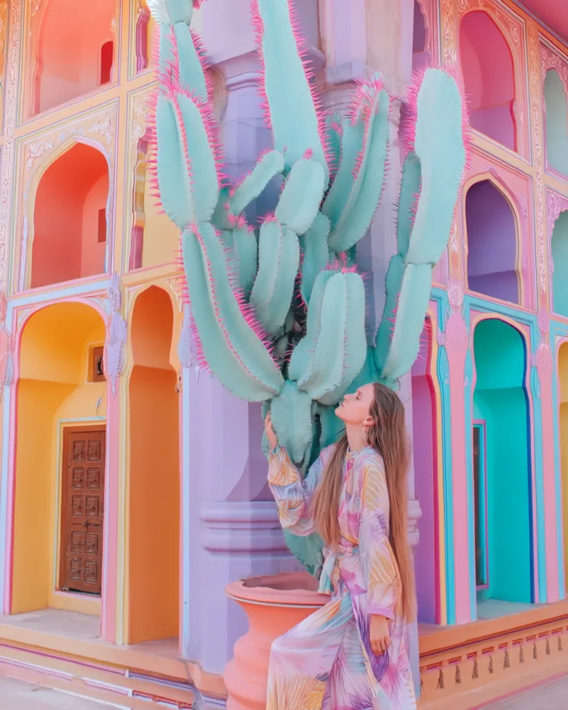 A woman with long blonde hair stands beside a large, vibrant green cactus with pink spines.  She's wearing a flowing pastel-colored dress. The backdrop is a brightly colored building with pastel orange, pink, purple, and teal walls, featuring intricate architectural details and arched doorways. The scene is whimsical and visually striking, a blend of nature and architecture.