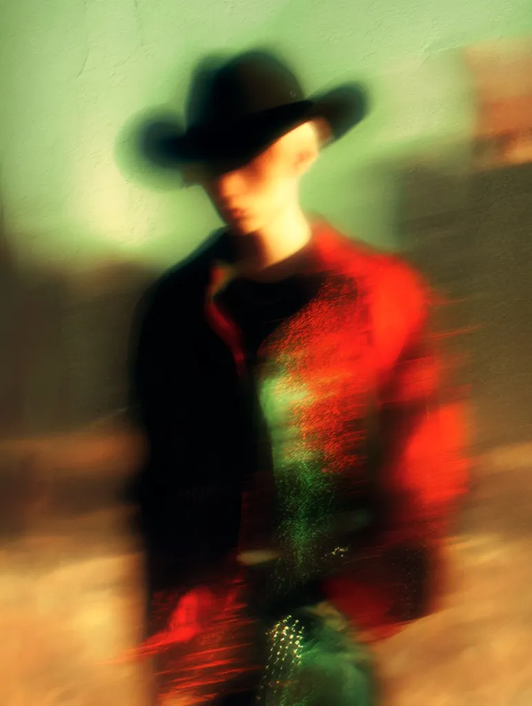 A blurry image depicts a person wearing a cowboy hat and a red and black jacket. The figure is slightly out of focus, creating a dreamlike or ethereal effect.  The background is a muted green, contrasting with the vibrant colors of the clothing. The overall impression is one of motion and mystery. The image's style suggests a deliberate artistic choice to blur the details, emphasizing mood over clarity.