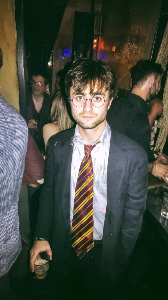 A young man, resembling Harry Potter, stands in a dimly lit bar. He wears round glasses, a dark suit, and a Gryffindor-colored tie.  He holds a beer, and other people are visible in the background, blurred by the low light. The overall atmosphere is casual and slightly dark. The image has a slightly grainy, vintage quality.