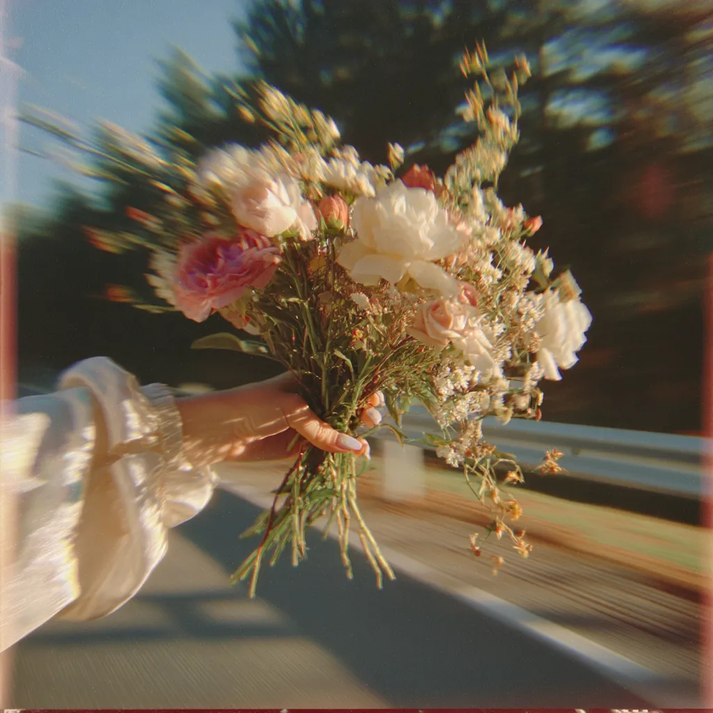 A hand holds a vibrant bouquet of pastel-colored flowers, predominantly white and pink roses and delicate greenery, against a blurred background of a sun-drenched road. The image has a dreamy, vintage aesthetic, with a soft focus and light leak effect, suggesting movement and a feeling of freedom and joy.  The overall mood is romantic and whimsical.