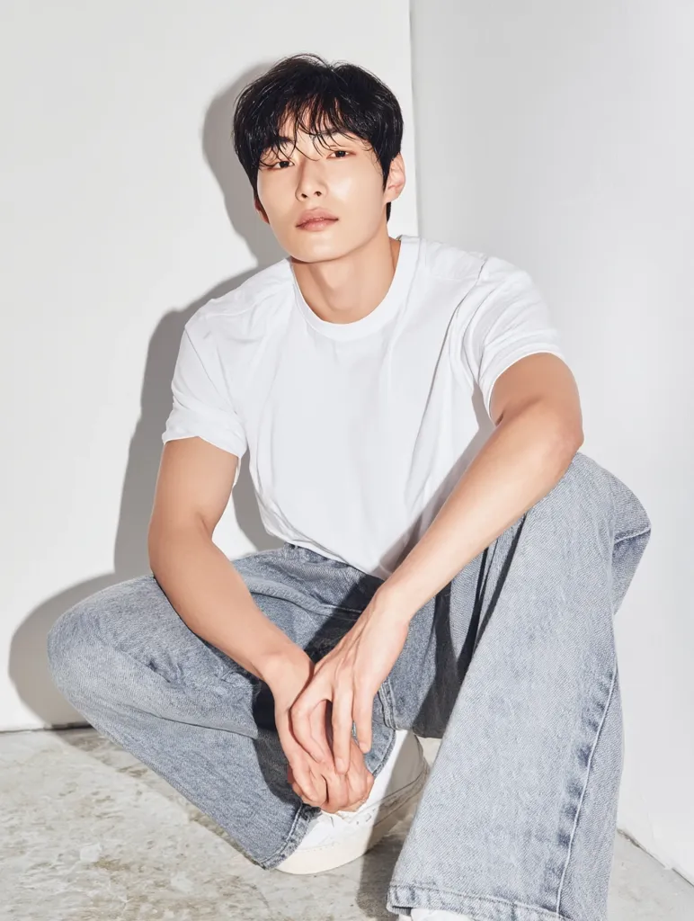 A young man with dark hair sits crouched on a light-colored floor against a white wall. He's wearing a simple white t-shirt and light gray, wide-legged jeans. His pose is relaxed, hands resting on his knees, and his gaze is directed toward the camera with a calm expression. The overall aesthetic is clean and minimalist.