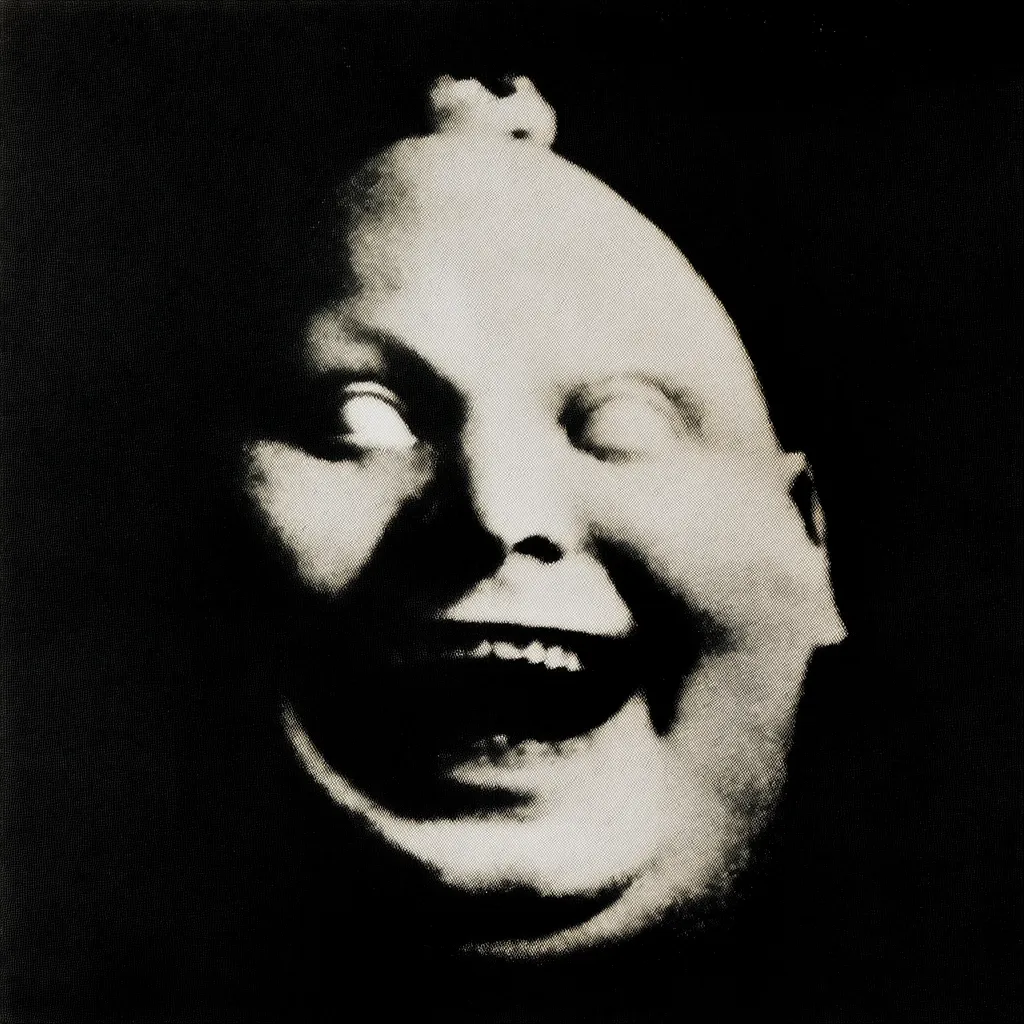 A black and white close-up photograph depicts a distorted, grotesque face. The subject appears to be a bald, chubby individual with exaggerated features.  Their mouth is open in a wide, unsettling grin, revealing teeth. The eyes are slightly rolled back, adding to the disturbing, surreal quality of the image. The overall effect is unsettling and evokes a feeling of unease.
