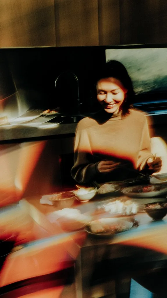 A young woman with dark hair sits at a kitchen table, illuminated by warm sunlight. She's wearing a light brown sweater and smiles gently as she seems to be preparing or eating a meal. The table is set with several small dishes containing food, creating a cozy and intimate atmosphere. The image is slightly blurred, adding to its dreamy and warm feeling.