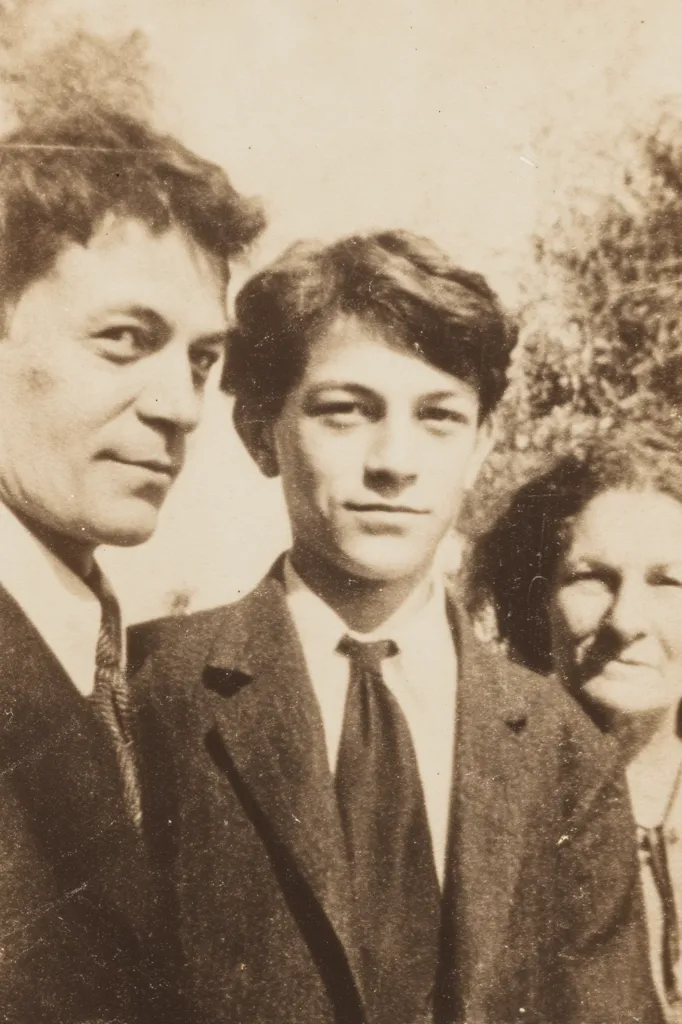 A sepia-toned photograph shows three individuals, likely a family.  A man in a suit and tie stands on the left, beside a younger man also in a suit and tie.  A woman with dark hair is partially visible on the right. The photo appears to have been taken outdoors, with some foliage visible in the background. The overall style suggests an early to mid-20th century setting.