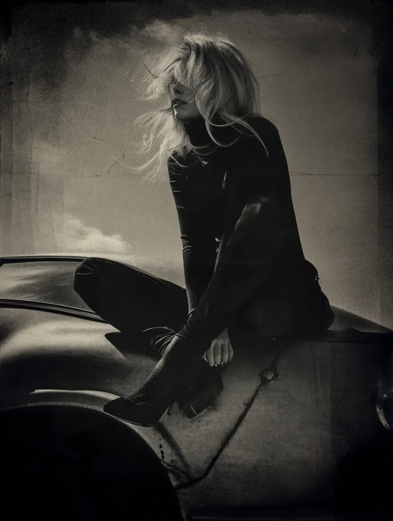 A monochrome image captures a woman with windblown blonde hair, seated on the hood of a weathered car.  She's dressed in dark, form-fitting attire, including tall boots.  The overall mood is melancholic and evocative, with the textured background adding to the image's gritty atmosphere. The contrast between the woman's soft hair and the harshness of the car creates a compelling visual narrative.