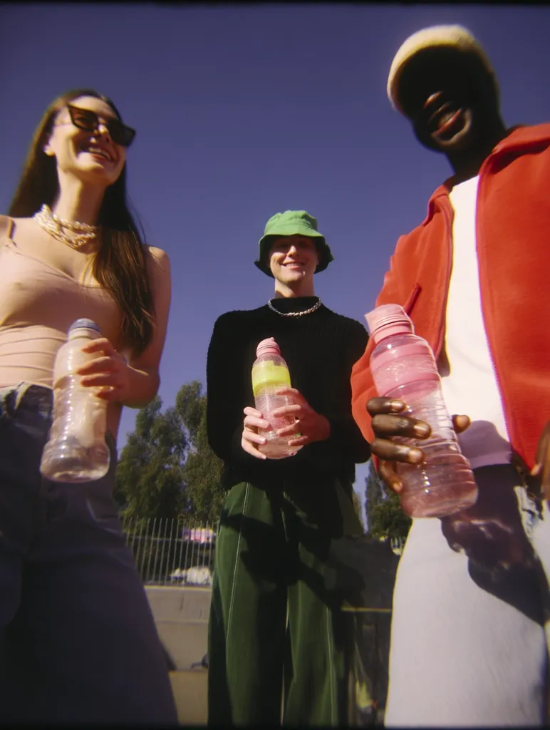 Three young adults, a woman with sunglasses and a beige top, a person in a green bucket hat and dark green pants, and a person in a red jacket, stand together outdoors on a sunny day.  Each holds a clear water bottle.  They look directly at the camera, creating a cheerful, vibrant scene. The background shows trees and a fence.