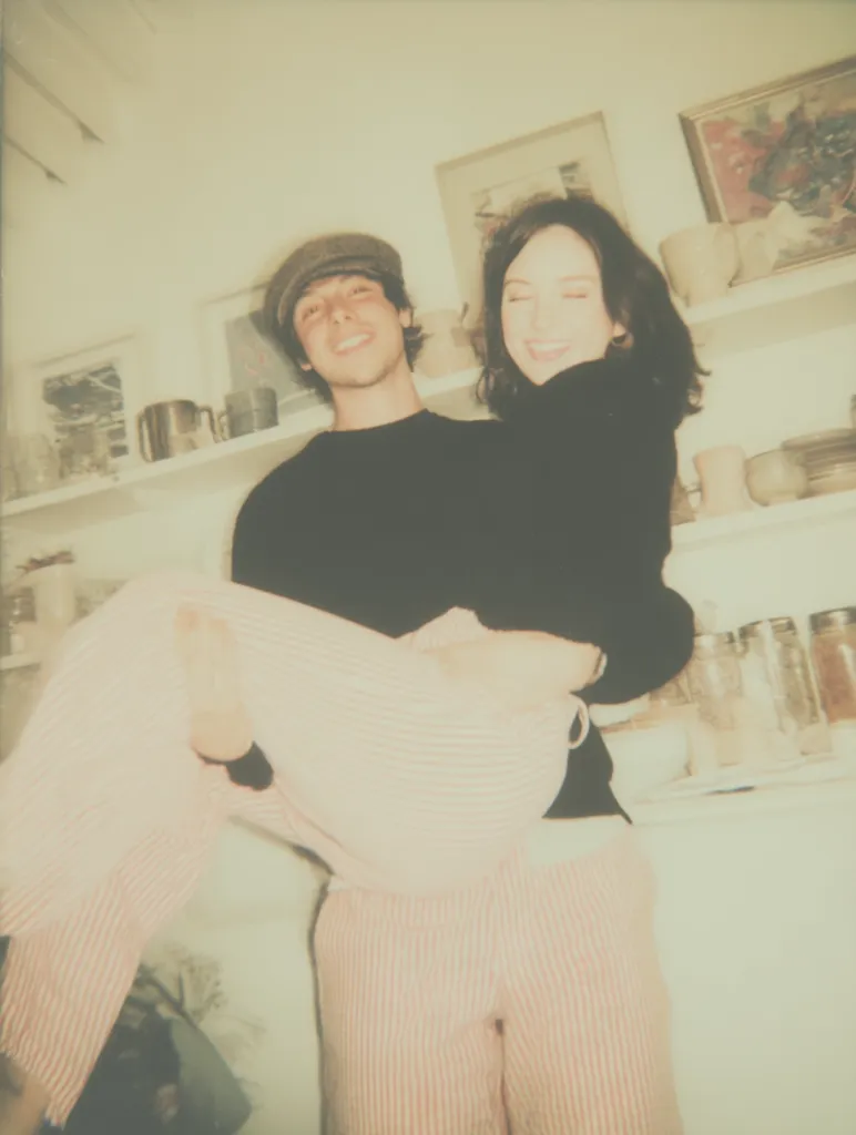 A man in a newsboy cap playfully carries a woman in pink striped pants.  The woman smiles, her dark hair framing her face.  They are in a kitchen with shelves holding dishes and jars, giving the photo a candid, intimate feel.  The image has a warm, slightly faded quality, reminiscent of a vintage photograph.