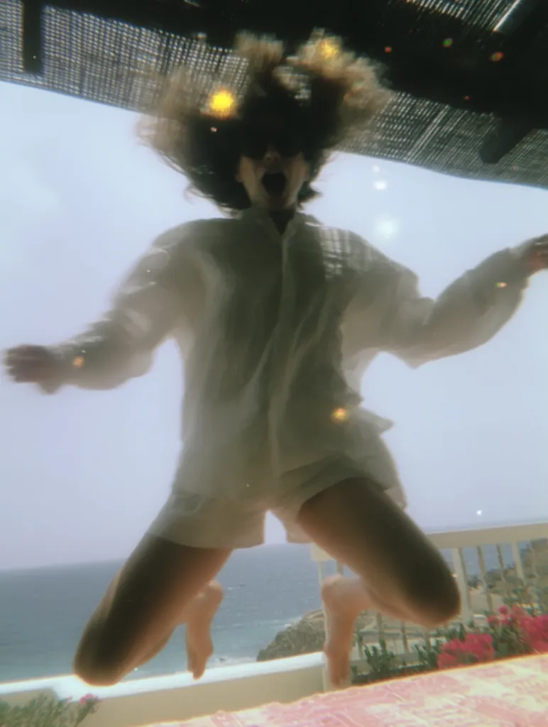 A woman in a white shirt and shorts joyfully leaps, her hair flying, against a blurred backdrop of an ocean view from a balcony.  The image has a dreamy, slightly out-of-focus quality, suggesting a carefree, spontaneous moment.  Sunlight filters through an overhead structure, adding to the ethereal atmosphere.