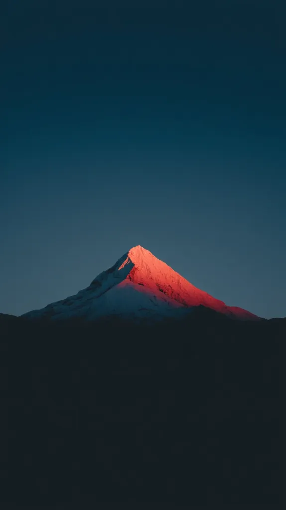 A snow-capped mountain peak is dramatically lit by the setting sun, casting a warm, reddish glow on its slopes. The sky above is a deep, twilight blue, providing a stark contrast to the vibrant mountain.  The lower portion of the image is shrouded in shadow, emphasizing the mountain's prominence against the darkening sky.  The scene evokes a sense of serene, majestic beauty.