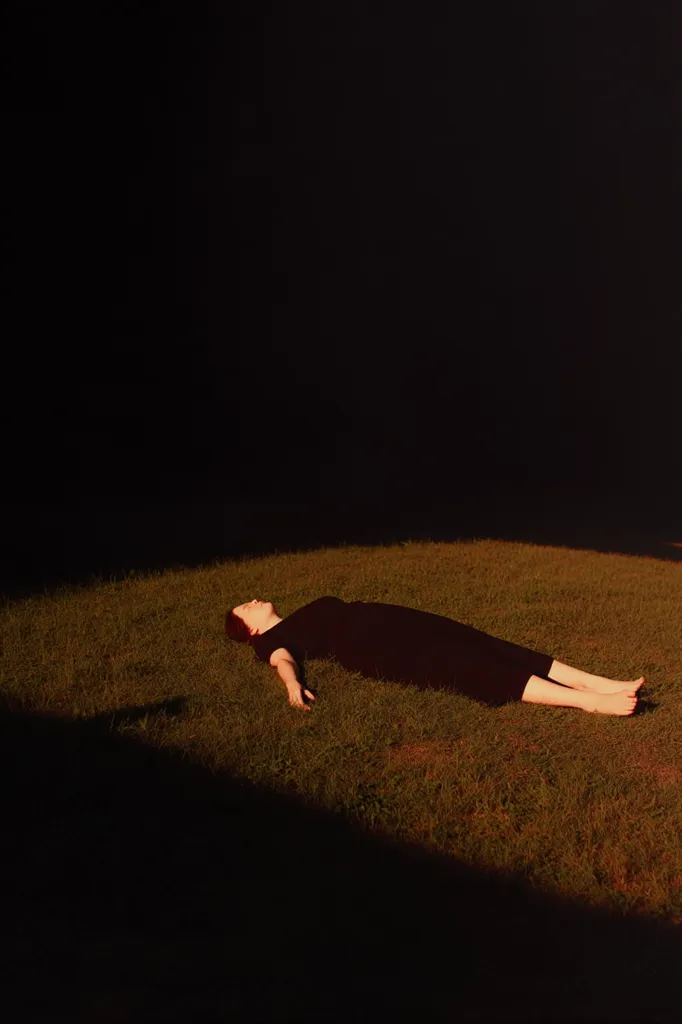 A person lies supine on short grass, arms outstretched, bathed in the warm glow of late afternoon sun.  Their dark clothing contrasts sharply with the golden light.  A deep shadow stretches from a dark background across the lower left of the image, creating a dramatic contrast with the illuminated figure. The scene evokes a sense of peace and solitude.