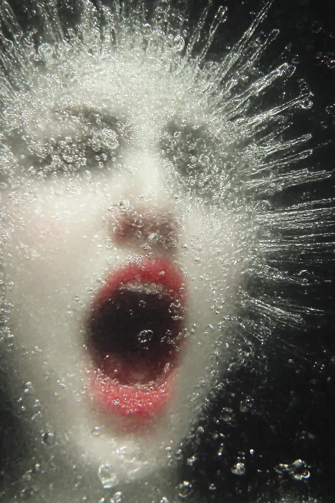 A close-up shot depicts a face submerged in water, seemingly screaming.  The water explodes outwards in a burst of droplets and streams, creating a dramatic, almost surreal effect. The face is partially obscured by the water, emphasizing the powerful emotion of the scream. The overall image is stark, with a dark background highlighting the bright, splashing water.