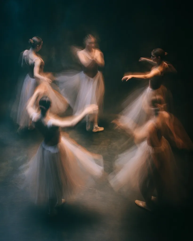 Five ballerinas in white tutus are captured in motion blur, their graceful movements creating a dreamy effect.  The dark background emphasizes the lightness of their costumes and the energy of their performance.  The image evokes a sense of fluidity and ethereal beauty, suggesting the intense effort and artistry of ballet.