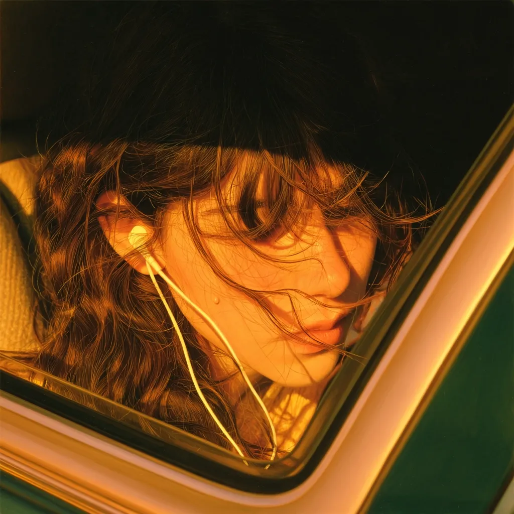 Bathed in golden sunlight, a young woman with long, wavy brown hair sits in a car.  Her face is partially obscured by her windblown bangs, and she wears white earbuds, suggesting she's listening to music. The warm, hazy light creates a nostalgic and dreamy atmosphere. The image focuses on her serene expression and the contrast between her hair and the car's interior.