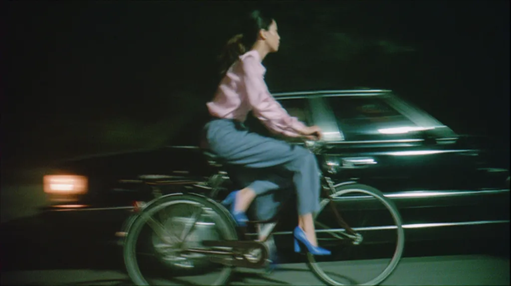 A young woman with dark hair, wearing a pink blouse and blue pants, rides a bicycle past a blurred, dark-colored car at night.  The scene is dimly lit, emphasizing the woman and her bike against the dark background.  The car is in motion, creating a sense of speed and movement. The overall tone is moody and atmospheric.