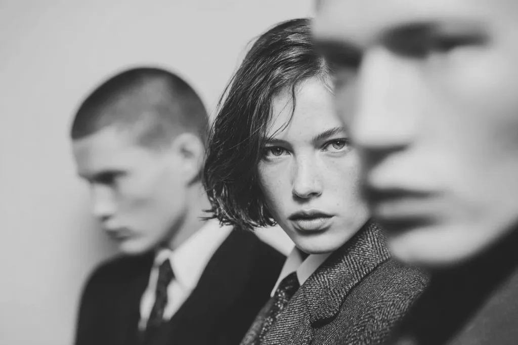 A black and white, close-up shot features a young woman with short hair, wearing a tweed jacket and tie.  She is in sharp focus, while two men in suits are blurred in the background, partially obscuring her.  The image conveys a sense of mystery and style, with a strong contrast between the woman's direct gaze and the men's out-of-focus presence.
