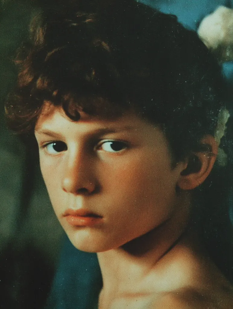 A close-up portrait shows a fair-skinned child with dark curly hair, possibly a boy. He faces forward, his gaze direct. The lighting creates a contrast, highlighting his features. His expression is thoughtful or serious, with slightly parted lips. The background appears to be a muted blue and green. The image quality suggests an older photograph or painting, with a slightly grainy texture. There is a decorative white floral detail behind his head on the right side of the frame.