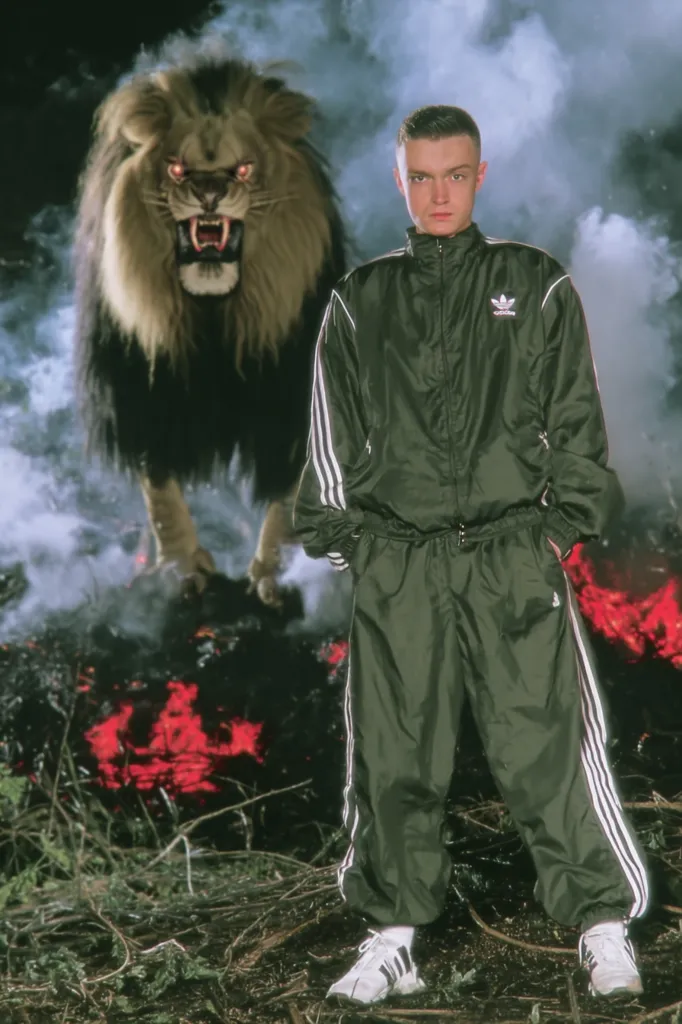 A young man in a full Adidas tracksuit stands before a roaring fire, a large lion looming behind him.  The scene is smoky and dramatic, with the red flames contrasting against the dark, nighttime setting. The man's expression is serious, his gaze directed forward. The overall mood is intense and somewhat mysterious.